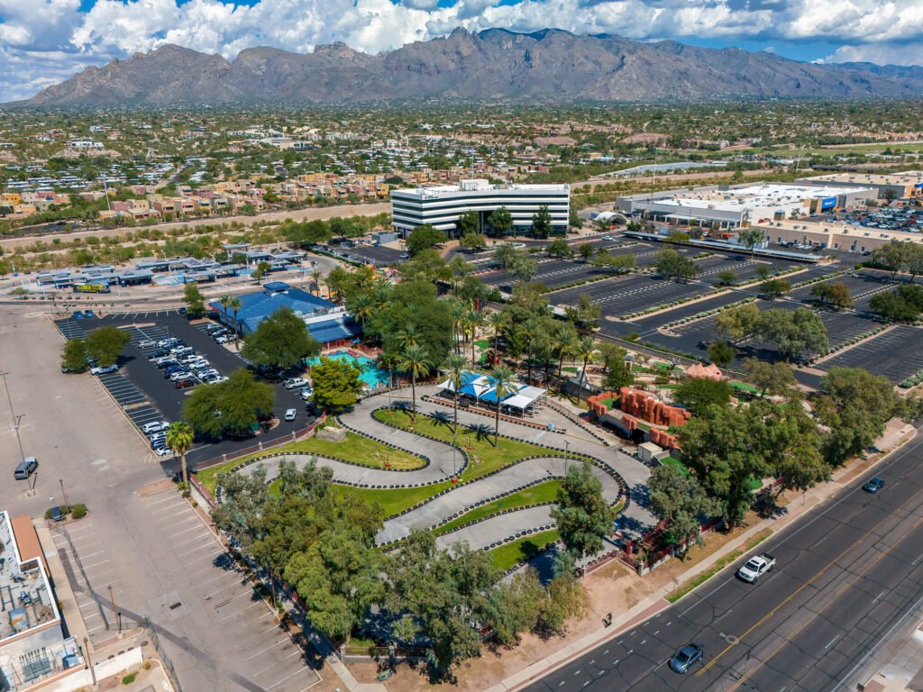 High-angle aerial drone photograph capturing the Tucson landscape, showcasing a go-kart track, large commercial parking lots, a white office building, and the Santa Catalina Mountains under a partly cloudy sky.